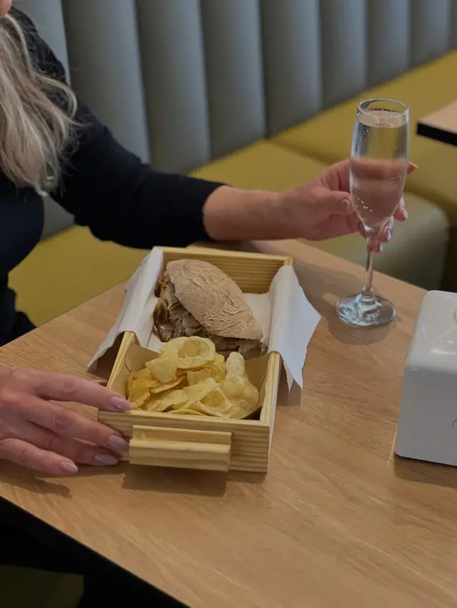 Cliente à mesa com uma sandes de leitão e batatas fritas numa caixa de madeira e um copo de espumante